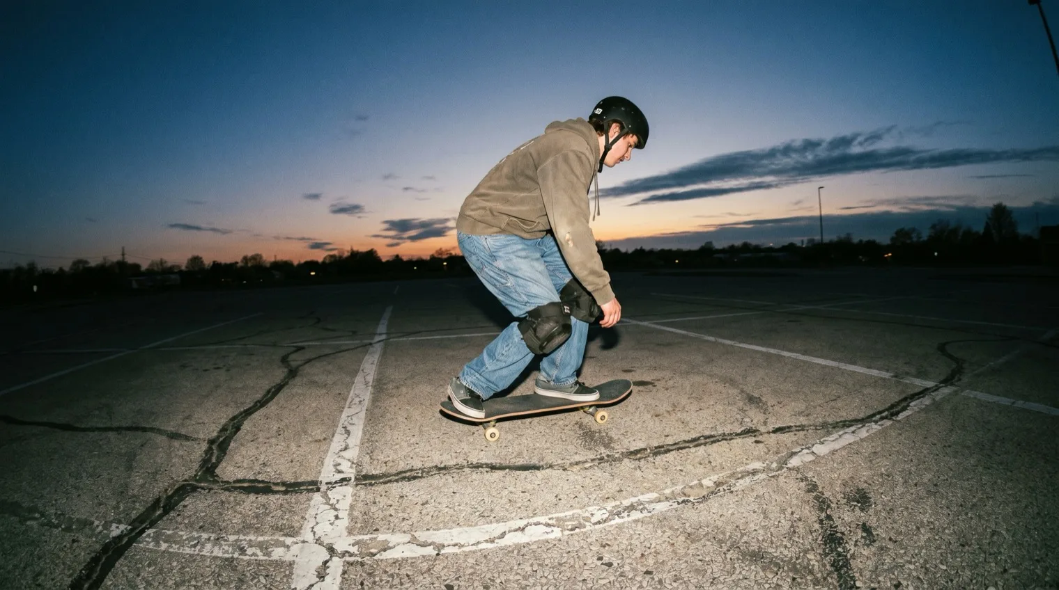 Beginner skater with helmet and pads rolling across a parking lot at dusk